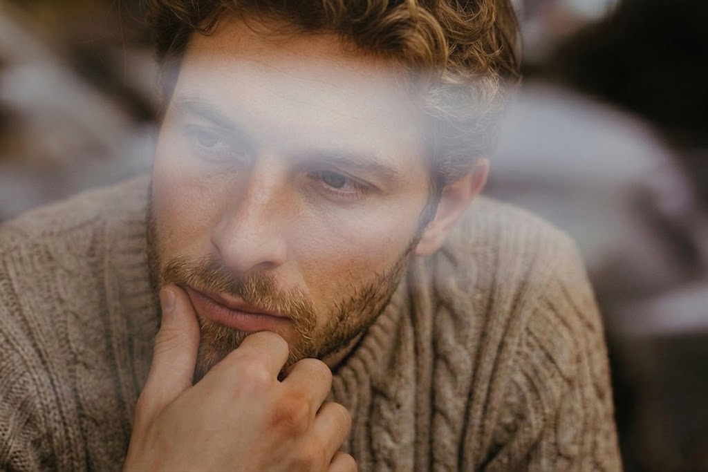 A man looking through a glass surface with his reflection visible, hand on chin in a thoughtful pose, representing the layered and partially hidden nature of subconscious patterns