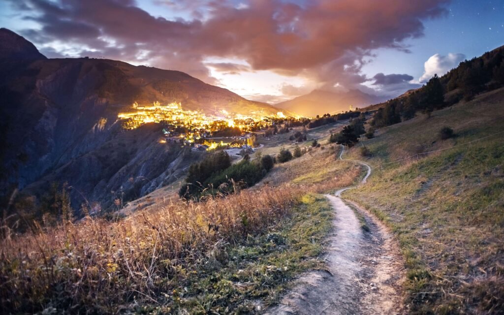 A winding mountain path leading down toward a warmly lit village at dusk, symbolising the journey ahead and the step required to begin it.